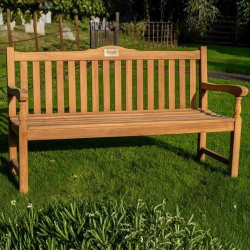 The 1.5m Teak Memorial Garden Bench by Bingley Fencing, featuring a slatted backrest and armrests, is set on a green lawn among trees and plants. A "Regal" plaque on the backrest adds style to this elegant outdoor seating option.