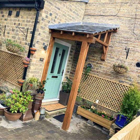 A house entrance features the Bingley Fencing Corner Oak Porch with mint green door, sturdy timber posts, and slanted roof. Potted plants, wall trellises, hanging baskets, and stone brick walls decorate the entryway.