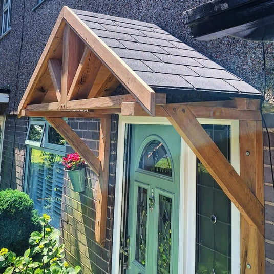 The Bingley Fencing Entrance Porch Canopy with a shingled roof is installed above a light green front door, featuring exposed beams and a hanging flower basket on the brick wall beside the entrance.
