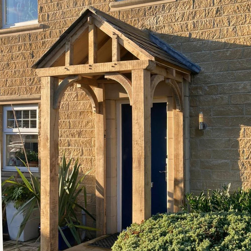 Load image into Gallery viewer, The Custom Made Oak Entrance Porch by Bingley Fencing features handcrafted joinery and a pitched roof over a dark blue door in a light stone house, with nearby greenery and sunlight casting shadows on the wall.