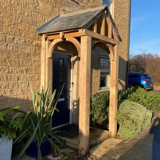 A modern house entrance features a dark blue front door, sheltered by the Custom Made Oak Entrance Porch from Bingley Fencing with handcrafted joinery and a slate roof. Potted plants line the doorway as sunlight falls on the stone walls.