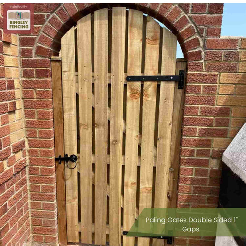 A Paling Gates Double Sided with 2" gaps (Flat Top & Arched Top) by Bingley Fencing is installed in a brick archway. The outdoor wooden gate features black metal hinges, a latch, and a small Bingley Fencing logo in the top left corner.