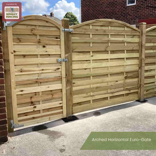 An Arched Horizontal Euro-Gate - AHG180 by KDM, featuring an arched timber top and metal latch, is installed on a concrete driveway beside brick buildings. A sign in the corner reads Installed by Bingley Fencing.