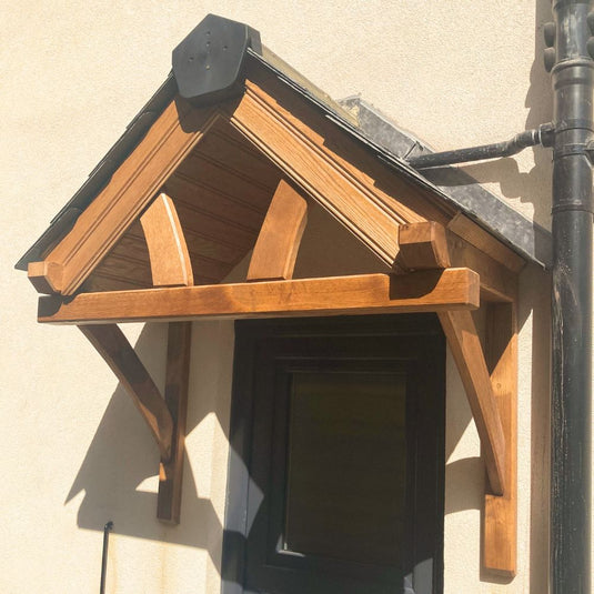 The Bingley Fencing Self Supporting Oak Canopy features a triangular roof above a black-framed door on a light exterior wall, supported by two wooden brackets and exposed beams.