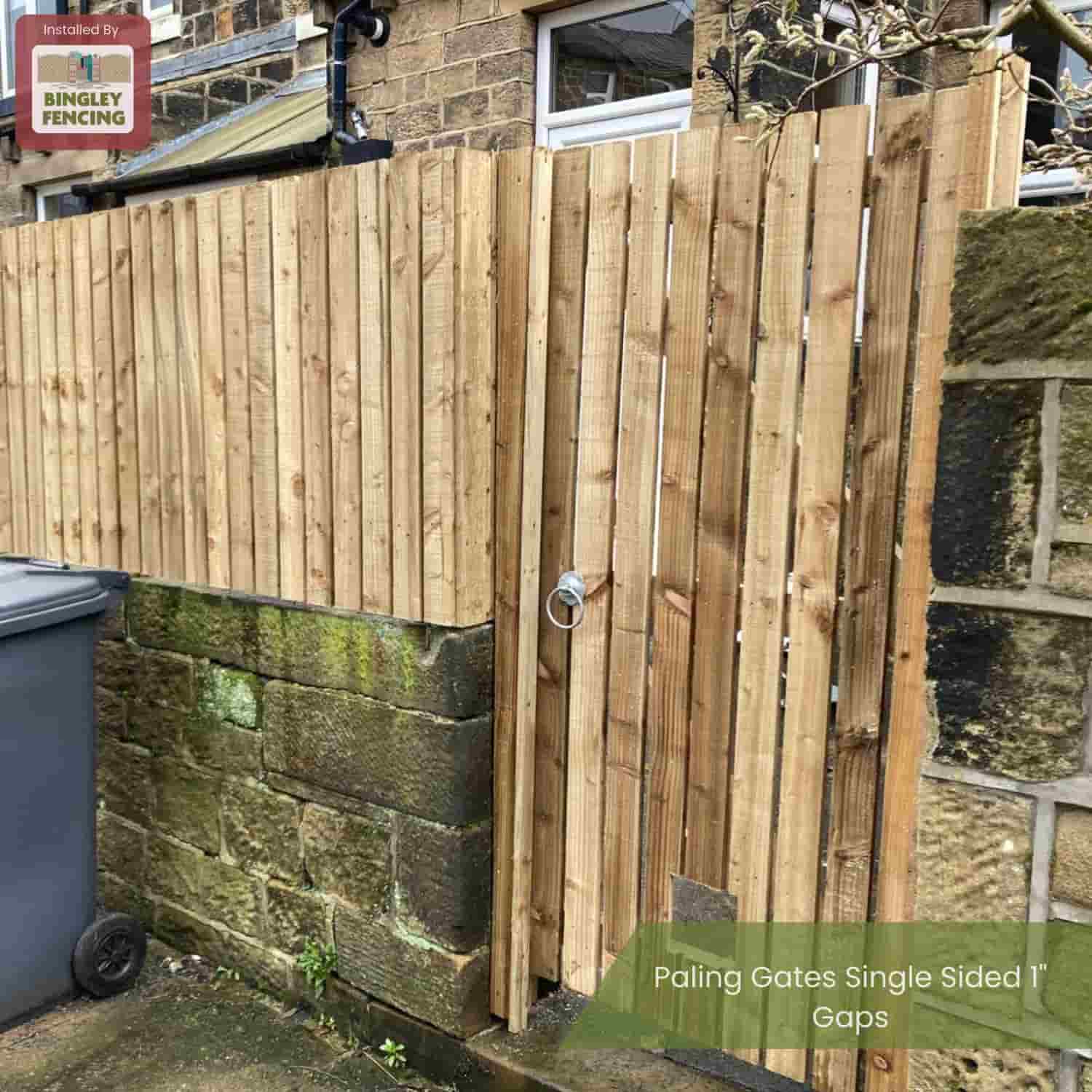 A Bingley Fencing Paling Gates Double Sided gate with 2" gaps and a flat or arched top, made from treated timber, is installed on a stone wall by a building. The outdoor gate features a metal ring handle, with a bin next to the wall.