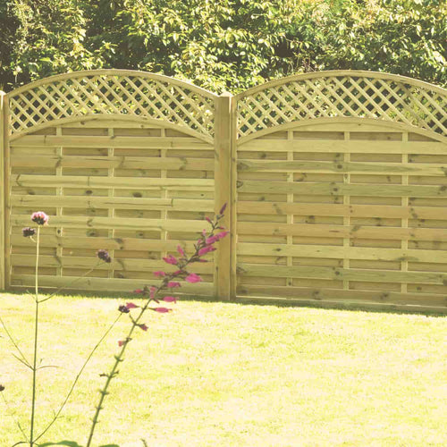 KDMs Arched Lattice Top Panels (ALT180) elegantly stand in a grassy yard with lush trees as a backdrop and pink-flowered branches enhancing the charm. Constructed from pressure-treated wood, these panels promise durability and beauty across seasons.