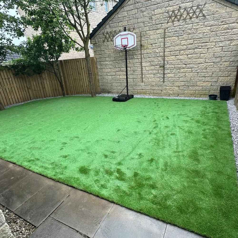 Load image into Gallery viewer, In the yard, the vibrant green turf from Bingley Fencing's "Artificial Grass Supply & Fit" enhances the space alongside a portable basketball hoop and brown stone walls. A tree graces one corner, while a wooden fence runs along one side. Gray stone tiles border the artificial grass, providing a seamless appearance thanks to the efficient installation service.