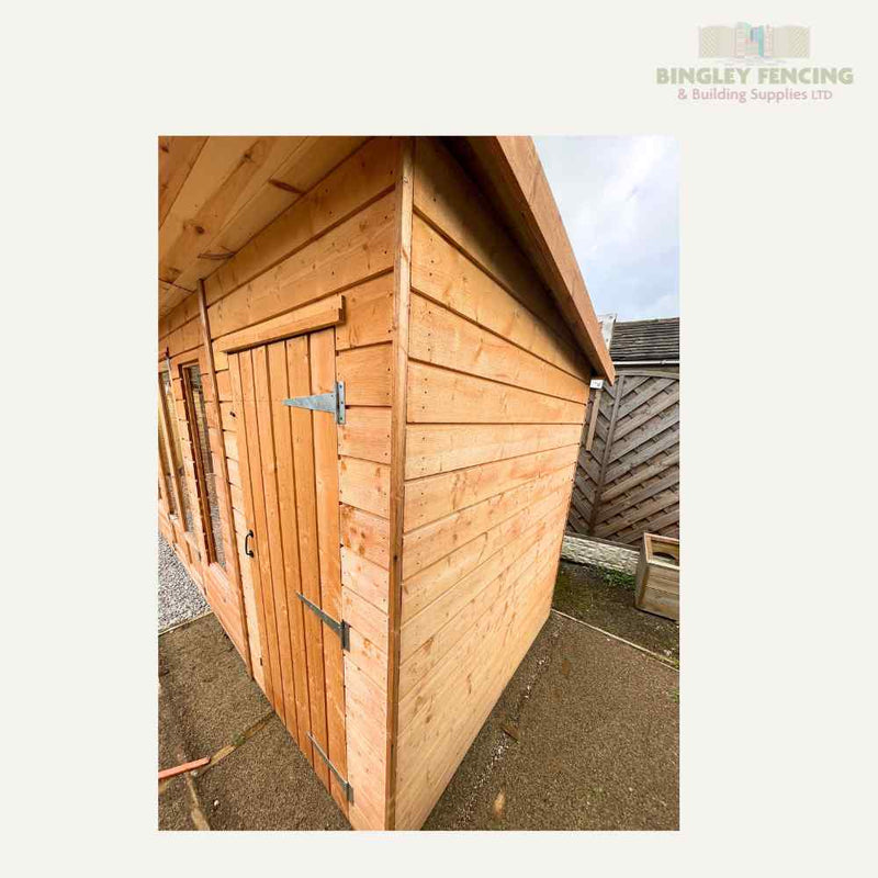 Load image into Gallery viewer, The Bingley Fencing Shed-Summerhouse Combination features custom-made vertical wooden panels, a concrete base, and a slightly sloped roof. It includes a door with metal hinges and latch, all set against a backdrop with fencing under cloudy skies.