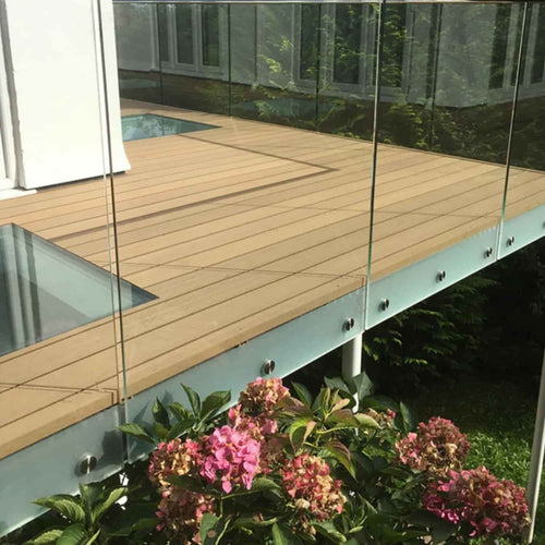 A glass railing with metal supports encloses a wooden deck built using Bingley Fencing’s deX Classic Composite Decking Boards 3.6m – 4 Colours, set above green bushes adorned with pink hydrangea flowers; modern building windows can be seen in the background.