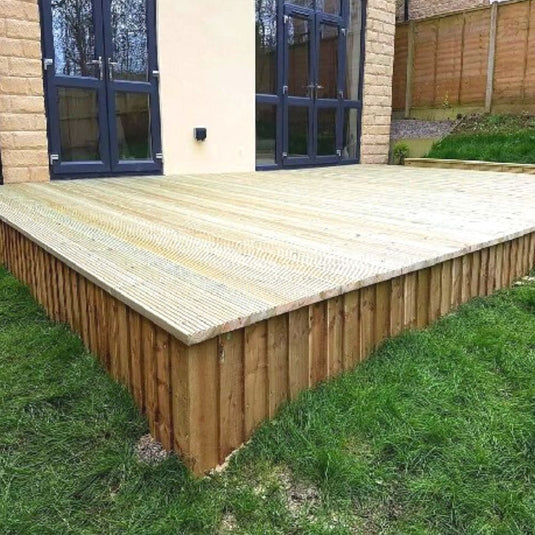 A newly built wooden deck is attached to the back of a house, bordered by green grass and a fenced yard, with large windows and doors leading inside.