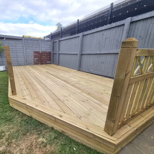 A newly built wooden deck with railings on one side, situated in a grassy backyard and surrounded by tall gray fencing under a partly cloudy sky.