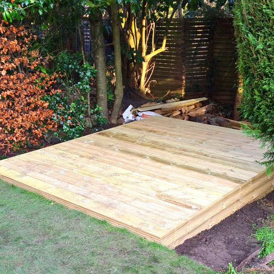 A newly built wooden deck sits in a garden corner, surrounded by grass, leafy trees, and shrubs, with some construction materials and tools visible in the background.