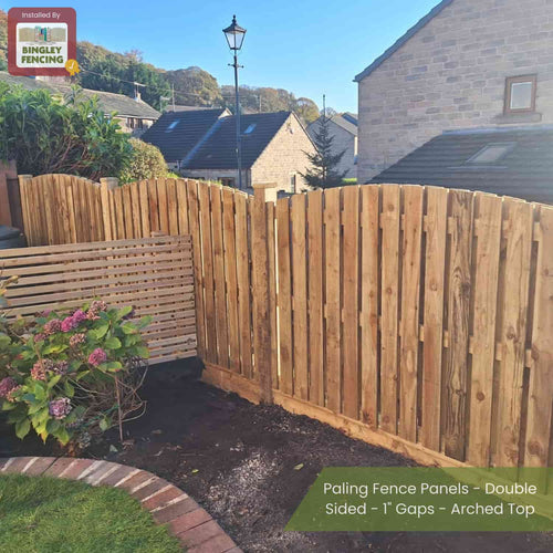 Bingley Fencing’s Double Sided Paling Fence Panels with 1 gaps and arched tops are installed in a sunny garden with houses behind; a sign reads Installed by Bingley Fencing. Flowering plants grow nearby.
