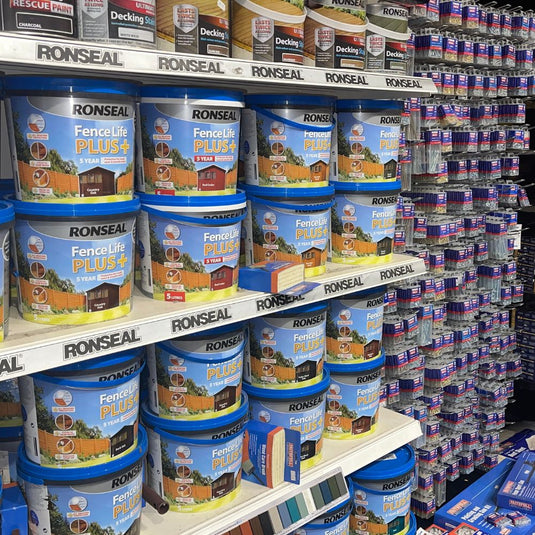 Rows of Ronseal Fence Life Plus paint tubs in blue containers are displayed on shelves in a store. To the right, shelves are stocked with various small boxed products, including Ronseal Rescue Paint.