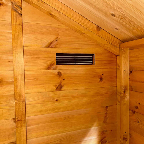 Interior view of a wooden wall showcasing the Bingley Fencing Shed Air Vent (Louvre Vent with Fly Screen) with horizontal slats installed near the top, below a sloped ceiling. Warm lighting highlights the natural wood grain.