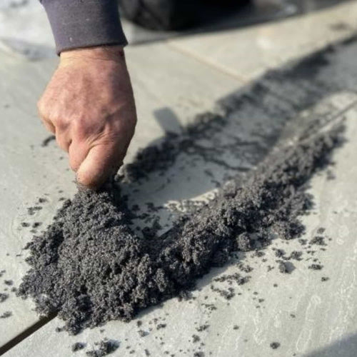 A hand spreads Joint-It Paving Jointing Compound 12.5kg by Joint It along the seams of light outdoor tiles, filling gaps and joints during installation on Indian sandstone.