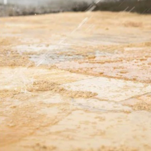 Close-up of water splashing over Indian sandstone joints filled with Joint-It Paving Jointing Compound 12.5kg by Joint It, highlighting the textured, muddy surface against a softly blurred background.