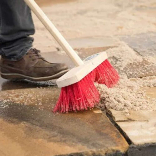 Wearing brown shoes, a person sweeps sand off Indian sandstone with a red-bristled broom, prepping the area for Joint-It Paving Jointing Compound 12.5kg by Joint It to create weed-resistant pavement joints.