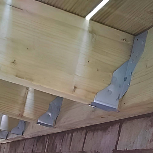Close-up of wooden beams secured to a brick or stone wall using Henry Shaw Long Leg Joist Hanger 50mm Timber 19" Long, viewed from beneath a deck or raised platform.