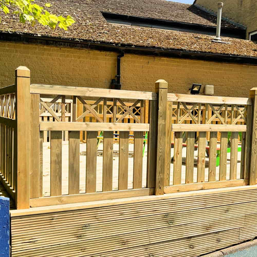 A wooden fence with decorative cross patterns encloses a sandy play area, set against a yellow brick building with a sloped roof on a sunny day.