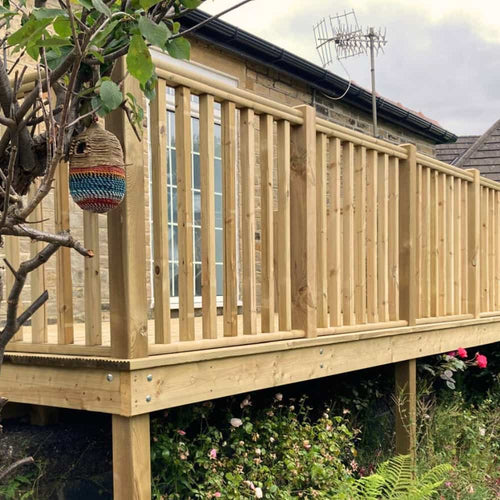 A wooden deck with vertical railing attached to a stone house, with a hanging bird feeder and flowering plants growing underneath. An antenna is visible on the roof in the background.