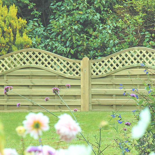 The KDM Omega Lattice Top Fence Panels, featuring a wavy top, stand elegantly in a garden with lush green foliage and vibrant pink and blue flowers. Constructed from pressure-treated wood, these panels ensure durability while adding European charm.