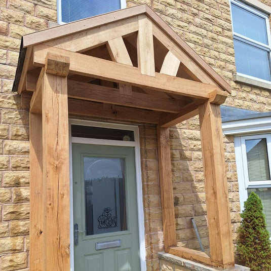 A light green front door with a decorative window sits in a stone wall beneath the Traditional Oak Porch & Frame by Bingley Fencing, featuring exposed beams, a triangular roof, and classic joinery for shaded, stylish entry.