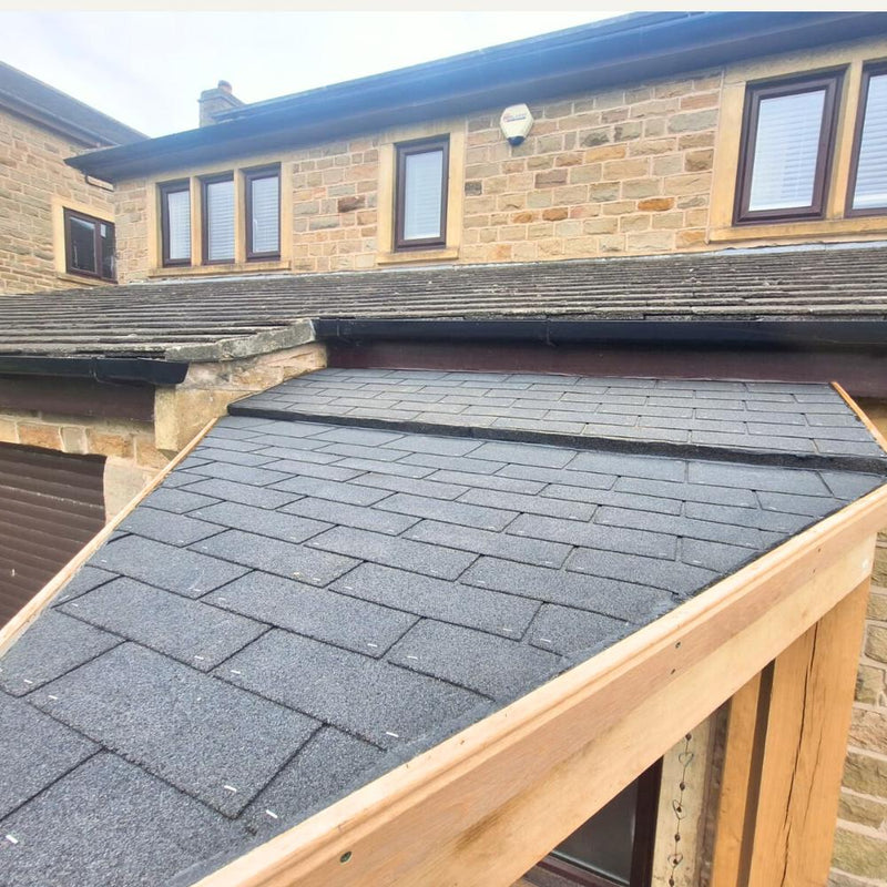 Load image into Gallery viewer, A close-up of a new shingle roof extension with black asphalt shingles, featuring the Oak Entrance Porch by Bingley Fencing next to a stone house with brown-framed windows and tan brick walls.