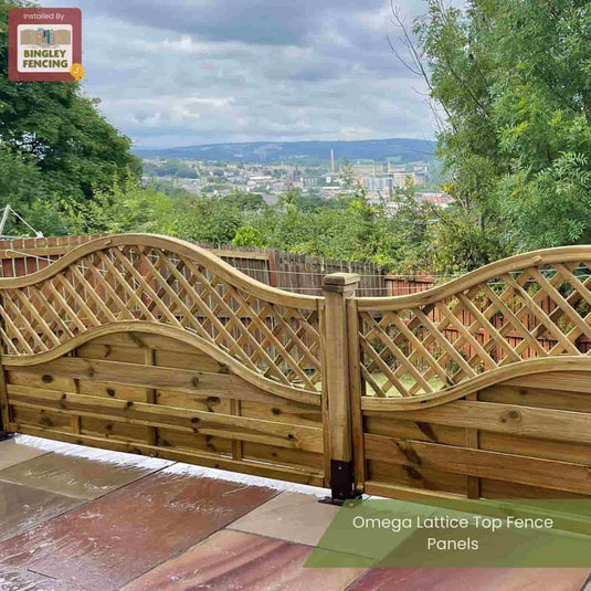 KDM Omega Lattice Top Fence Panels with a decorative curved lattice stand on a patio, overlooking a scenic town amid trees and hills. Text: KDM Omega Lattice Top Fence Panels – pressure treated for lasting beauty.