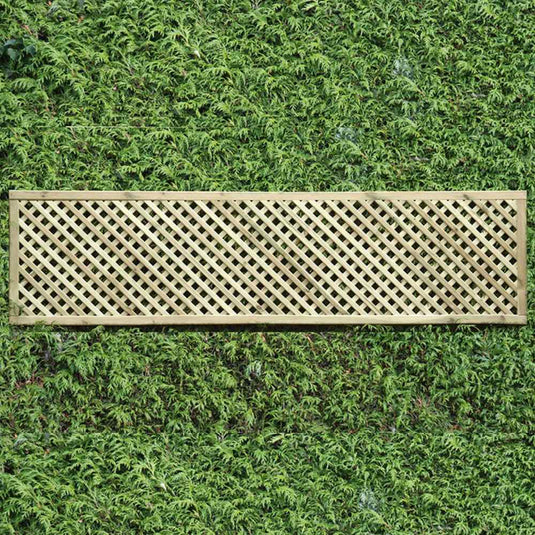 The KDM Trellis - Privacy Lattice - Rectangle 45cm x 183cm (PL8) is a pressure-treated wooden panel with diamond-shaped openings, laid horizontally against a green hedge for both charm and seclusion in your garden.