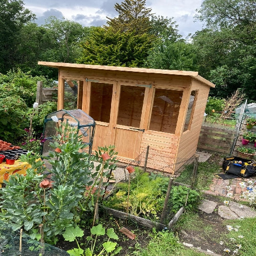 A small wooden shed with a flat roof stands in a lush garden. The garden features green plants and flowers, with stones and a path leading to the structure. There are trees in the background under a cloudy sky. A toolbox lies nearby.
