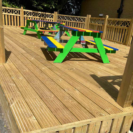 A wooden deck with colorful picnic tables featuring green legs and red, blue, and yellow tops. Each table has attached benches of matching colors. A wooden fence surrounds the deck. Trees and a building are visible in the background.