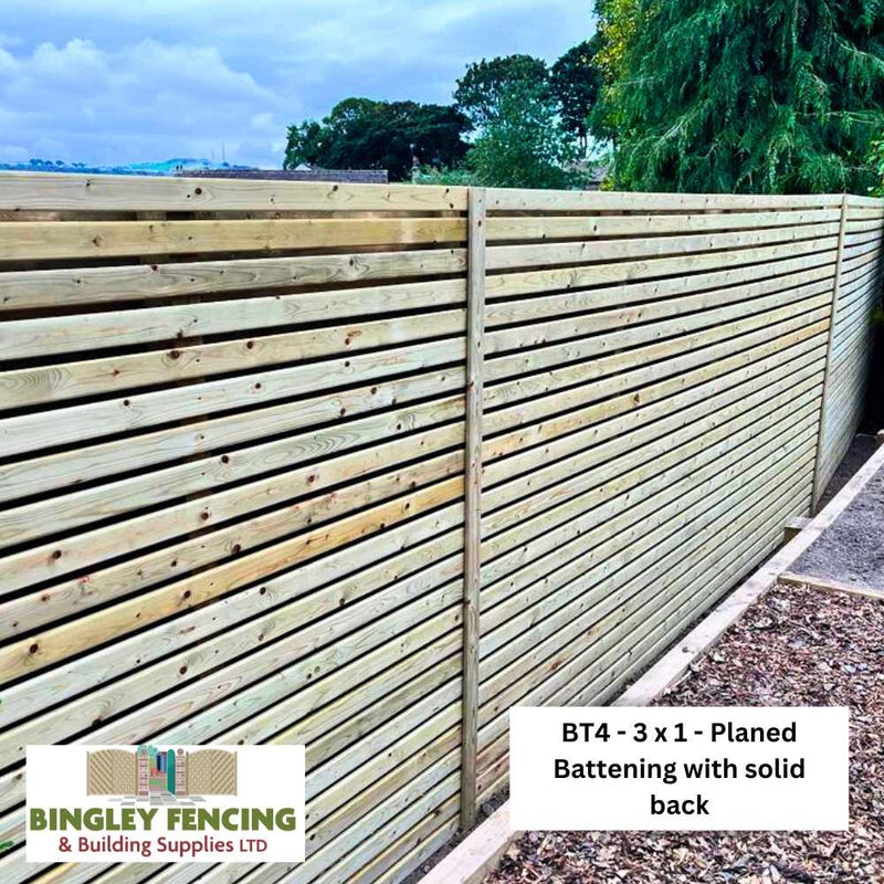 Load image into Gallery viewer, A tall wooden fence of Planed Round Edge Batten - 2” x 1” (50mm x 25mm) by KDM with a solid back stands beside a path. The KDM logo and a label with the product name appear in the image.