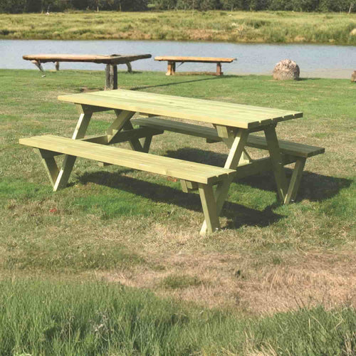 A KDM Premium A Frame Table 150cm x 180cm (PB180) with attached benches sits on grass by a pond, with timber garden products and European-style fence panels among trees and benches in the background under a clear sky.