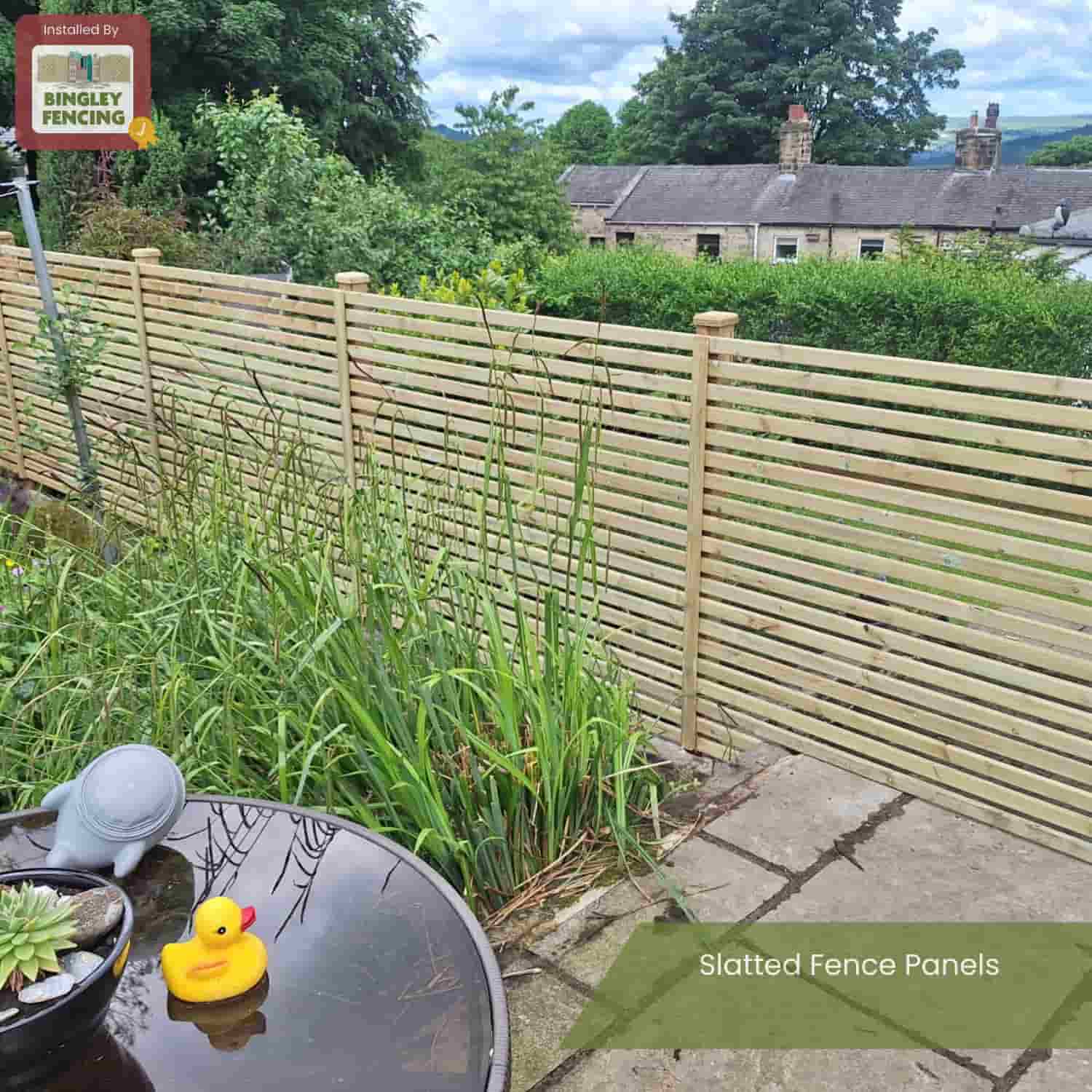 A garden with tall grass features a round table with a plant and yellow rubber duck, and a contemporary KDM Slatted Fence Panel in a light color. Trees and houses can be seen beyond. Text reads: Slatted Fence Panels.