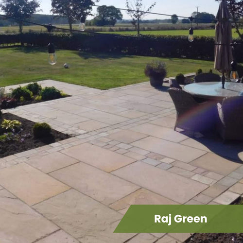 A natural stone patio with Bingley Fencing’s Indian Sandstone Paving Project Pack leads to a grassy yard edged with trees and shrubs. On the patio sits a round table with chairs and umbrella. Text overlay: “Raj Green.”.