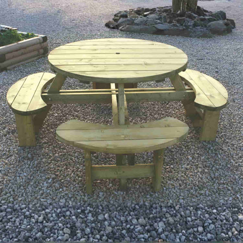 The KDM Round Table & Bench Seat - RBS features four attached curved seats, making it practical garden furniture for outdoor gravel areas with natural elements like trees and rocks in the background.