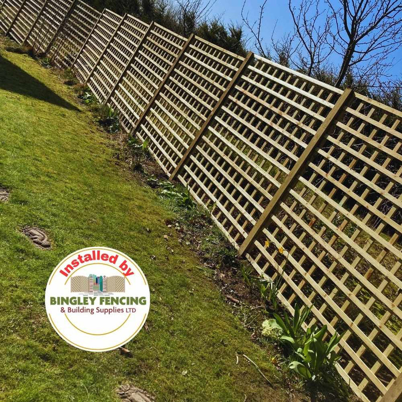 Load image into Gallery viewer, A row of Square Trellis Panels made from pressure-treated wood for added durability runs along a grassy garden with small plants at the base. A circular logo at the bottom reads, "Installed by Bingley Fencing.
