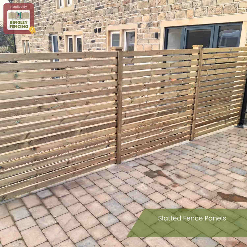 Load image into Gallery viewer, A KDM Slatted Fence Panel with a modern slatted design stands on a paved patio beside a stone building, offering partial privacy. It features a Slatted Fence Panels label and KDM branding in the top left corner.