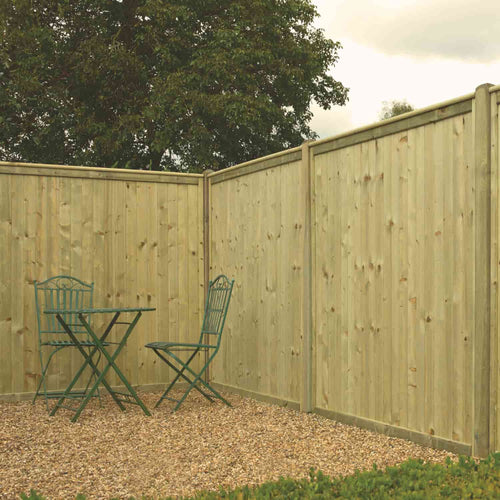 A compact outdoor seating area features a green metal table and two chairs on gravel, enclosed by tall KDM Tongue & Groove Fence Panels Flat Top (TGF), with a tree visible beyond the privacy fencing.