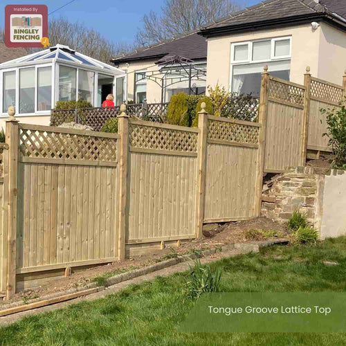 A KDM Reduced 5ft High Tongue & Groove Lattice Top Fence (TGLT150) runs along a sloped yard by a beige house and glass conservatory, bordered by green grass and a small garden.