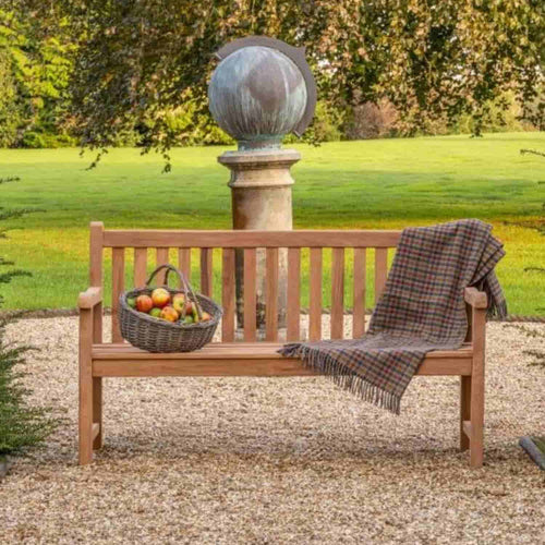 A 1.5m Teak Garden Bench by Bingley Fencing sits on a gravel path in a garden, accessorized with a plaid blanket, apple basket, and backed by a stone sphere on a pedestal and green lawn.