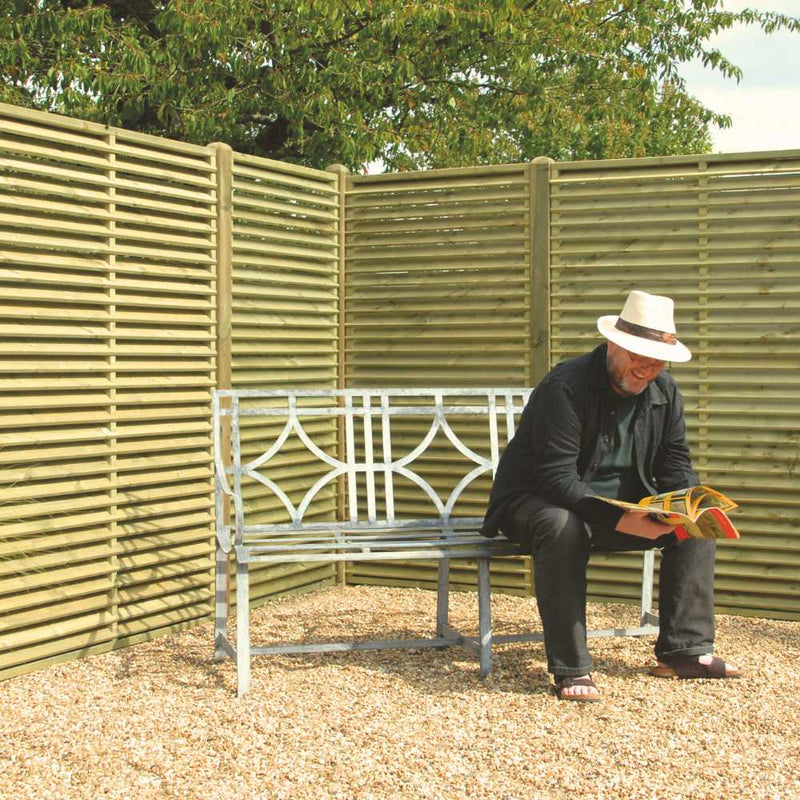 Load image into Gallery viewer, A man in a white hat and dark clothes reads a magazine on a metal bench in a gravel area bordered by KDM Reduced Venetian Fence Panels (VP180), with leafy tree branches visible above the privacy fencing.