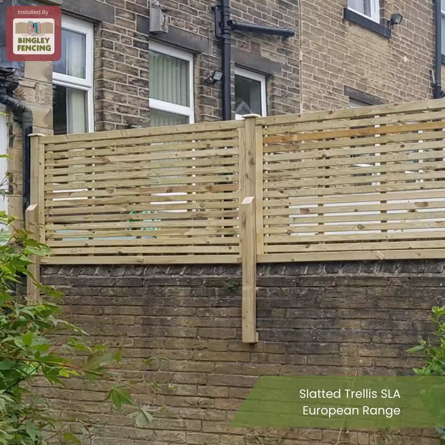 A slatted wooden trellis fence sits atop a stone wall outside a brick house with white-framed windows. The fence is from the European Range by Bingley Fencing. Green bushes are visible in the foreground.