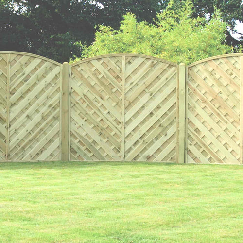 The KDM 'V' Arched Solid Fence Panel - VA features arched tops and a diagonal crisscross pattern, installed on a well-manicured lawn with lush green trees and bushes as a scenic backdrop.