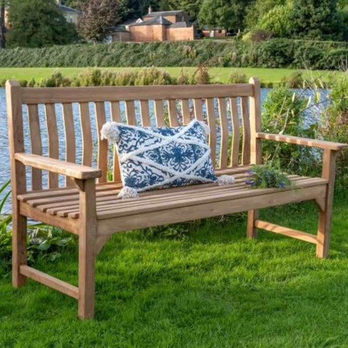 A 1.5m Wave Back Teak Garden Bench from Bingley Fencing, accented with a decorative blue and white pillow, sits on green grass beside a pond, providing charming outdoor seating with trees and houses in the background.