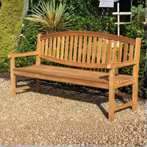 A 1.8m Curved Back Teak Garden Bench by Bingley Fencing, featuring a slatted backrest and armrests, sits on a gravel surface in a garden surrounded by green plants and shrubs.