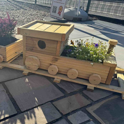 The KDM Hand Crafted Wooden Train Planter sits on a stone path filled with flowers, sunlight casting shadows. In the background, a sign with a QR code is visible.
