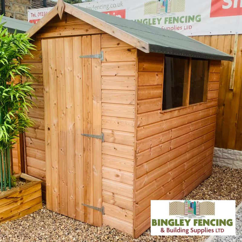 Load image into Gallery viewer, A wooden shed featuring an Apex Roof design and a single door is set against a gravel surface, with a bamboo plant positioned to the left. The Bingley Fencing logo is displayed in the bottom right corner, showcasing their sheds and customisation options. Company-branded signs can be seen in the background.