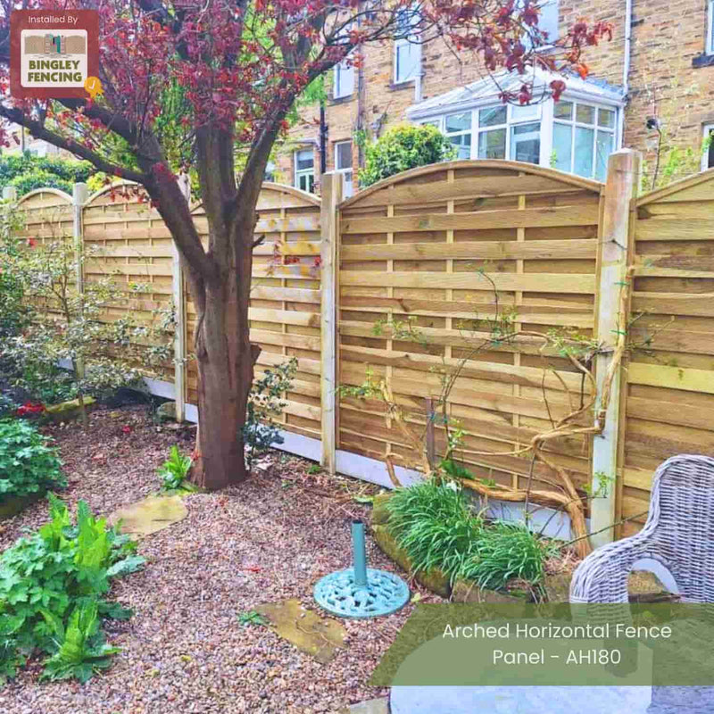 Load image into Gallery viewer, A garden with gravel paths, lush plants, a tree, a wicker chair, and tall KDM Arched Horizontal Fence Panels - AH180. Made from pressure-treated timber for elegance and durability.